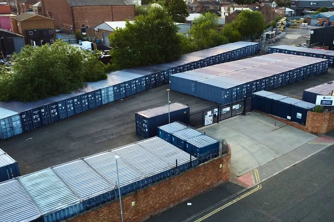 Aerial view of a container storage facility with multiple blue shipping containers arranged in rows, surrounded by brick walls and some trees.