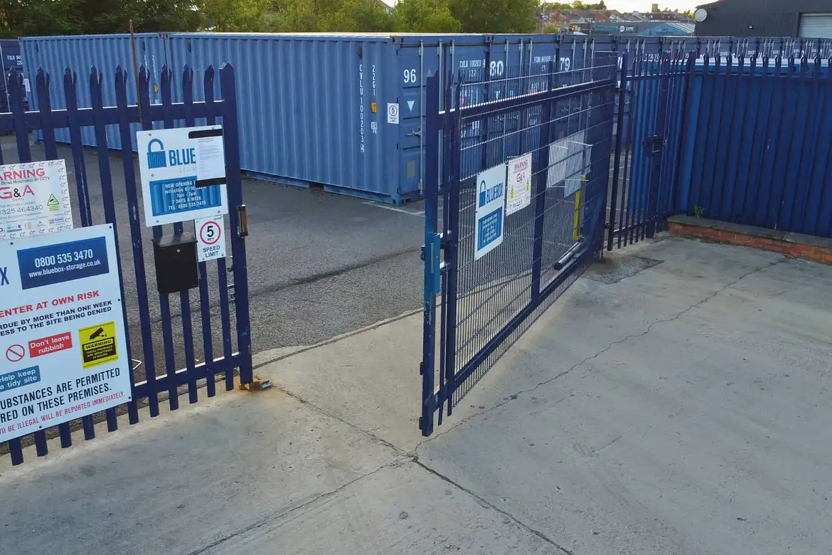 A blue metal security gate is partially open, revealing container storage units behind it; multiple warning and information signs are posted on the gate and fence.