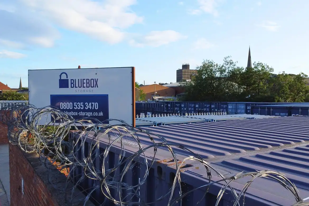 Rows of blue shipping containers behind a barbed-wire fence at Bluebox Storage, with a sign displaying the company’s name, phone number, and website—offering secure container storage. Trees and buildings appear in the background.