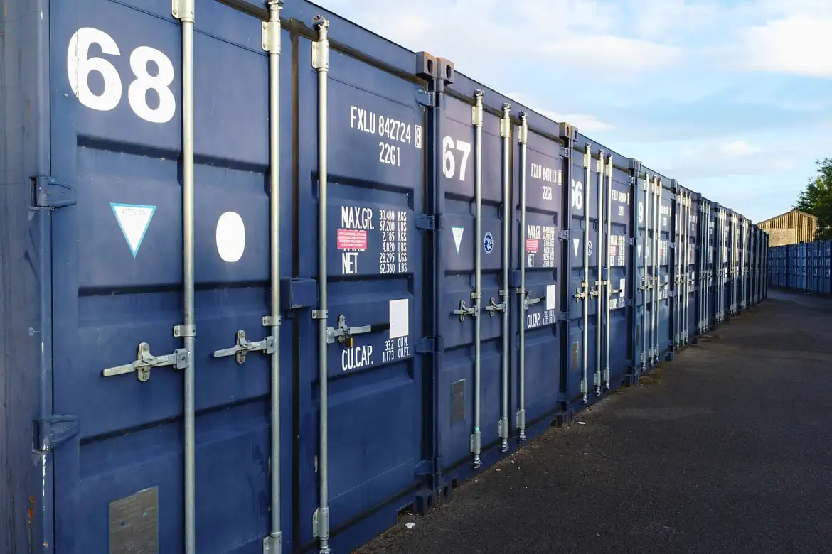 A row of blue container storage units with white stencilled numbers and metal locks, lined up on a tarmac surface under a partly cloudy sky.