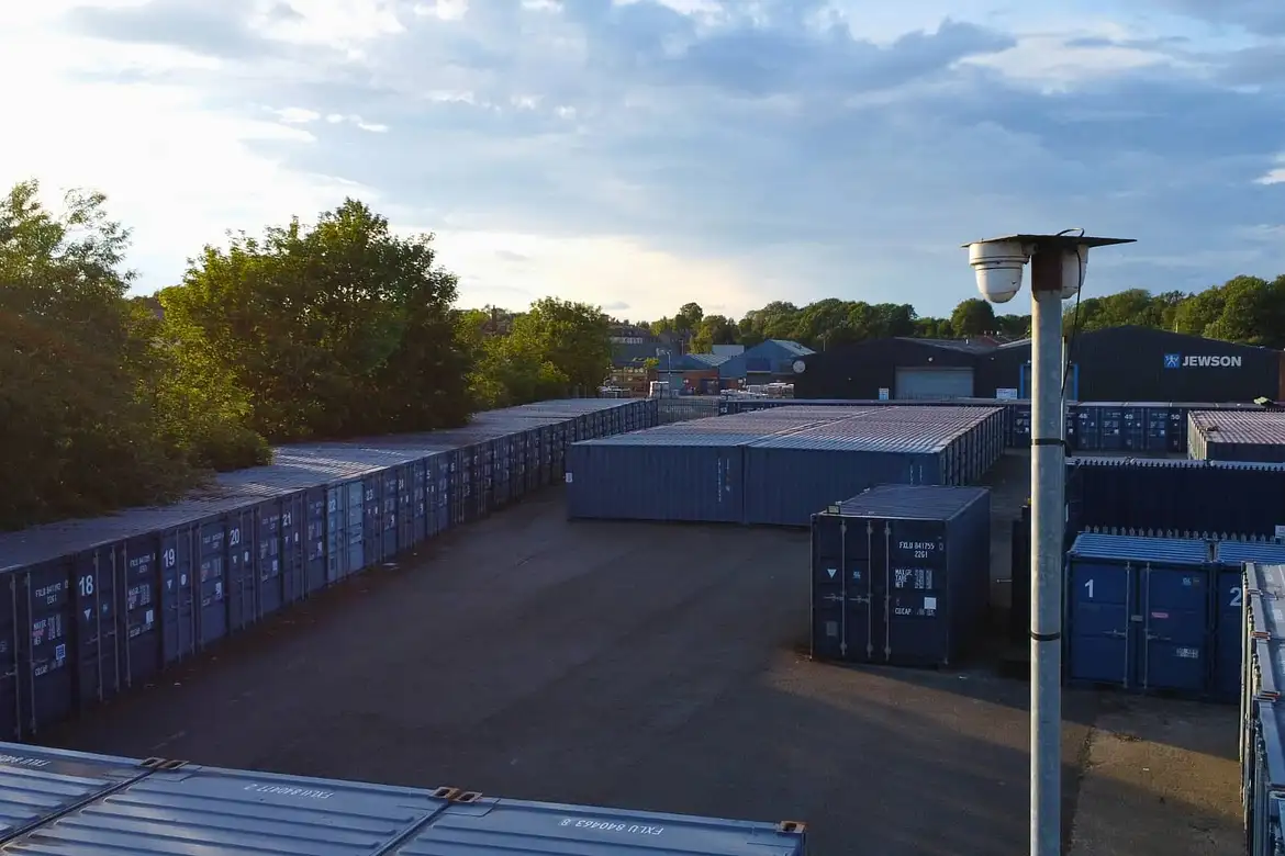 Rows of blue container storage units are arranged along a paved car park next to trees and industrial buildings, with a CCTV camera mounted on a tall pole in the foreground.