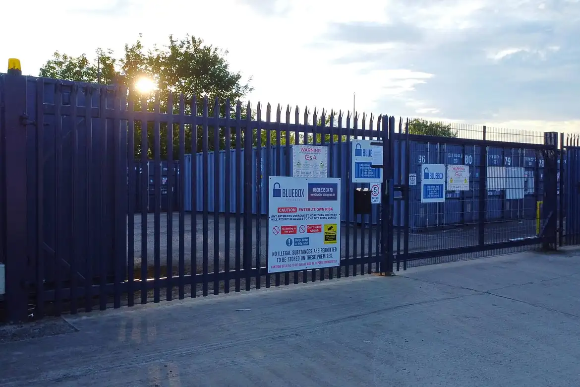 Secure self storage facility with blue metal fencing and gates, warning and business signs at the entrance, and container storage units visible inside. Sun sets behind trees in the background.