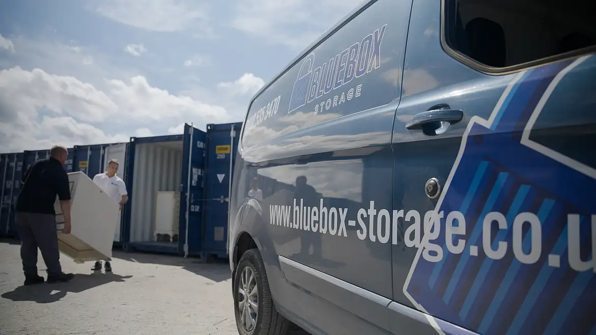 Bluebox Website-02 (1) Two people carry a large white object near a row of container storage units; a blue van with "bluebox-storage.co.uk" branding is parked nearby.