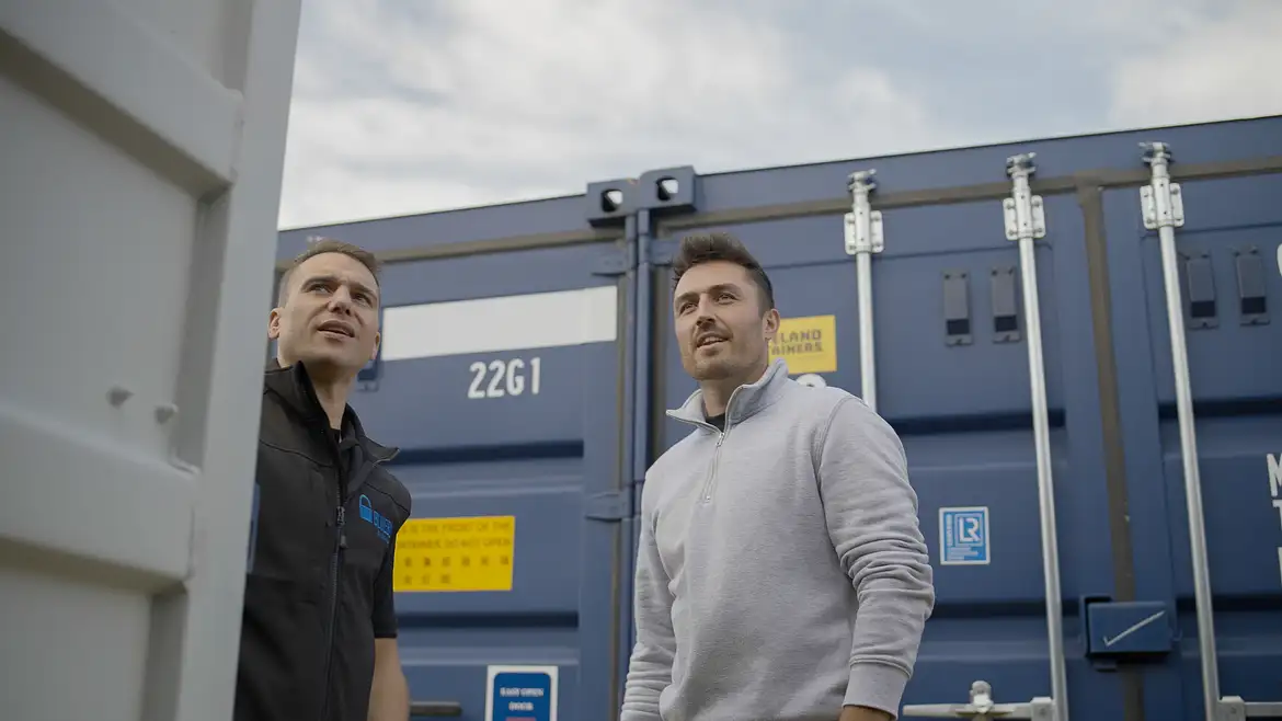 Two men stand outside, looking up, in front of a blue self storage shipping container with various labels and markings visible.